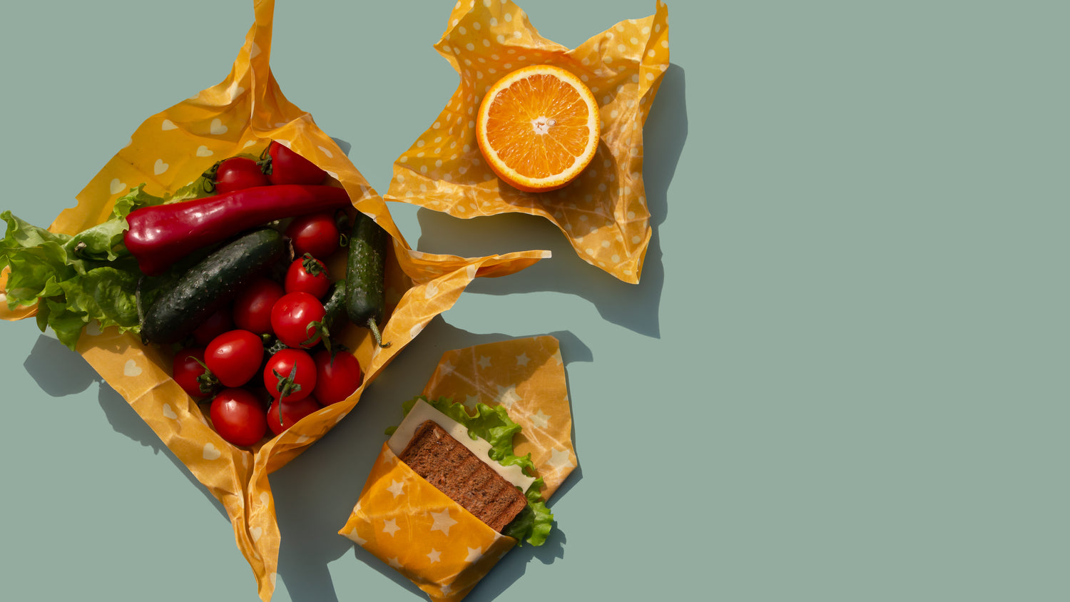 A variety of fresh produce and a sandwich wrapped in reusable beeswax food wraps on a pale green background